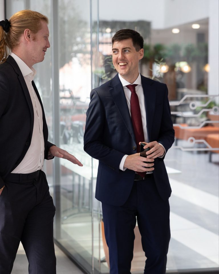 Two business colleagues talking in a modern glass-walled office lobby, on-location corporate photography by Leon Britton