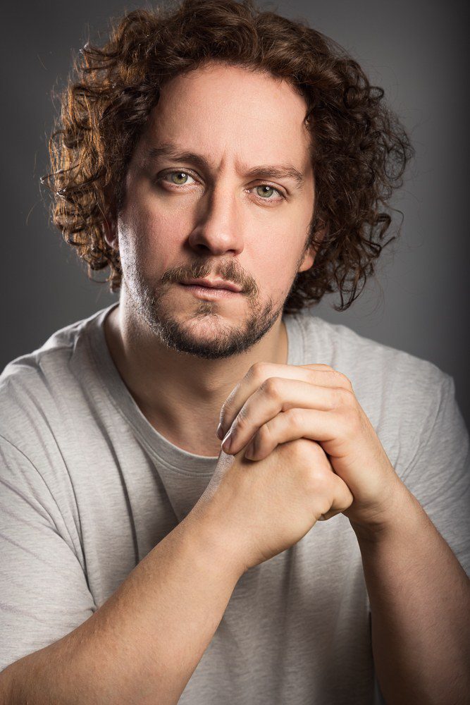 Adam Shaw, Liverpool actor, headshot portrait against a grey backdrop with hands clasped