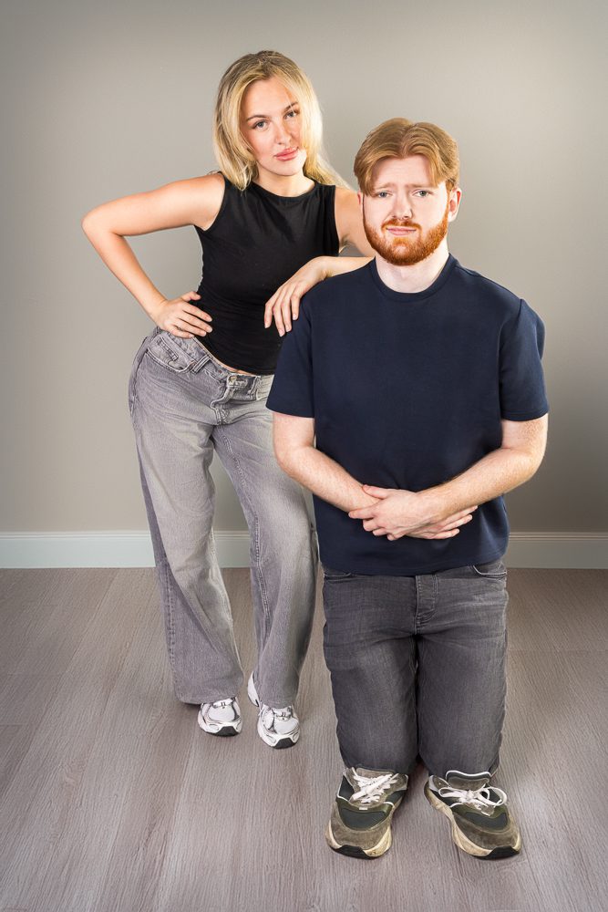 Woman standing confidently with hand on hip while her partner kneels awkwardly beside her in the studio