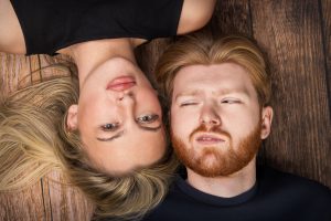 Couple lying head to head on wooden studio floor, one upside down, both pulling deadpan awkward faces