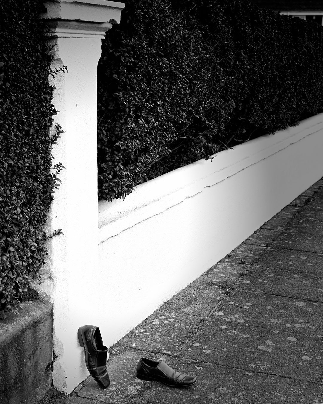 Black and white photograph of an empty pair of shoes against a garden wall and hedge