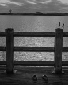 Black and white photograph of an empty pair of shoes left on a jetty overlooking water, conceptual photography project by Leon Britton